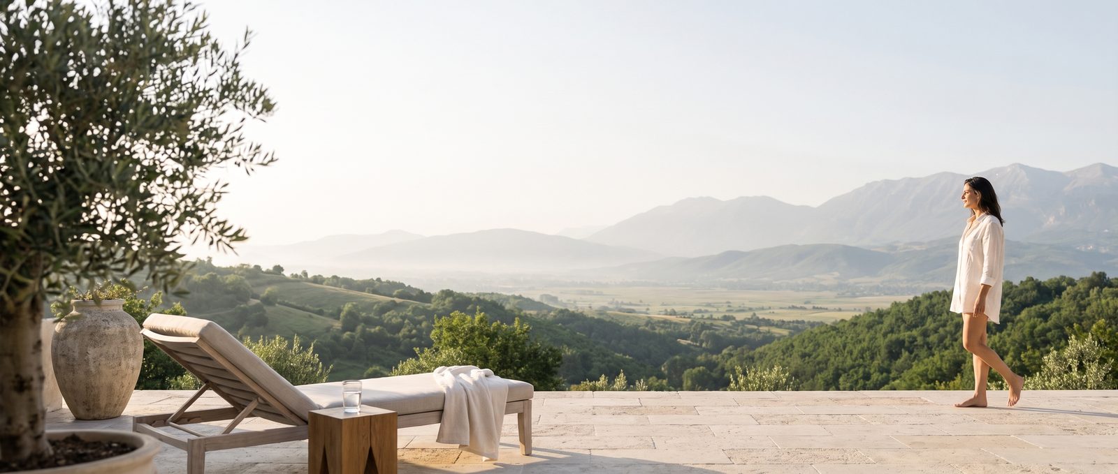 A calm hotel terrace in the morning, overlooking green hills and mountains, with a woman in a linen shirt walking toward the view.